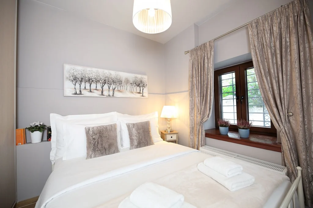 Υπνοδωμάτιο με διπλό κρεβάτι, απαλό φωτισμό από επιτραπέζιο φωτιστικό, και παράθυρο με κουρτίνες και δύο γλαστράκια στο περβάζι.