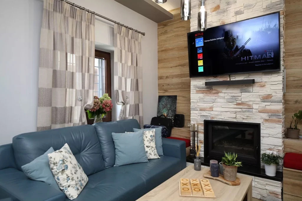 Ζεστό σαλόνι με μπλε καναπέ, διακοσμητικά μαξιλάρια, τηλεόραση με Netflix, τζάκι με πέτρινη επένδυση και φυσικό φωτισμό.
