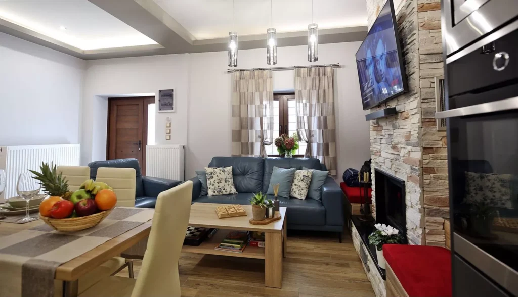 Σαλόνι μοντέρνου σπιτιού με μπλε καναπέδες, πέτρινο τζάκι, τραπέζι με φρούτα και τηλεόραση τοίχου.