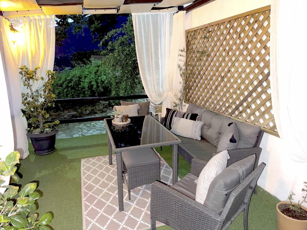 Εξωτερικό καθιστικό με καναπέ, τραπέζι και διακόσμηση σε αυλή με φυτά και κουρτίνες